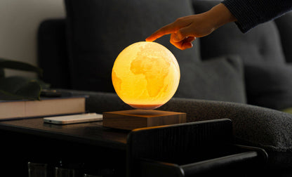 A hand touching the Gingko Smart Earth lamp, the lit globe floating above its wooden base