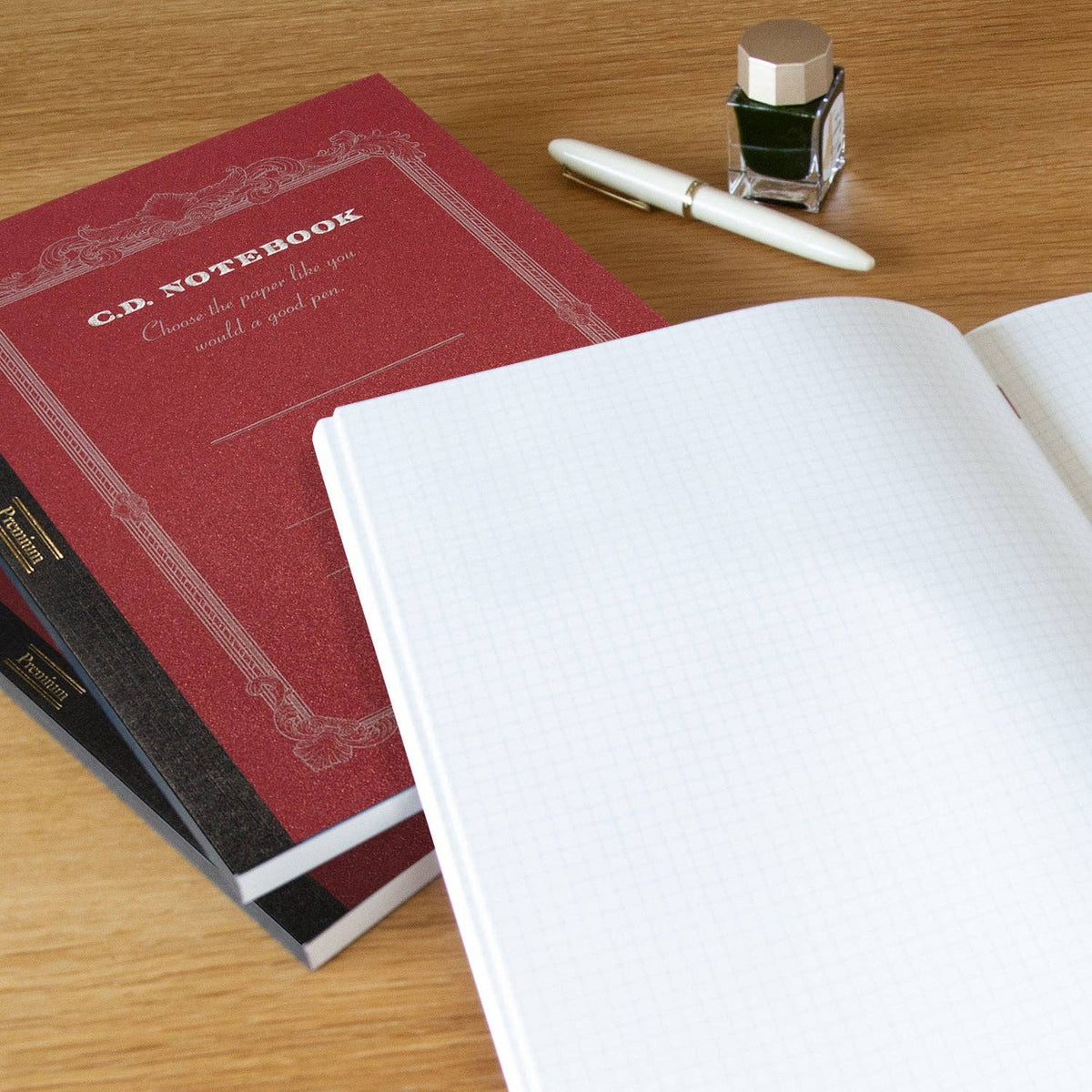Apica Premium A5 notebook in red open flat to grid pages, shown with a fountain pen and ink bottle on a wooden desk