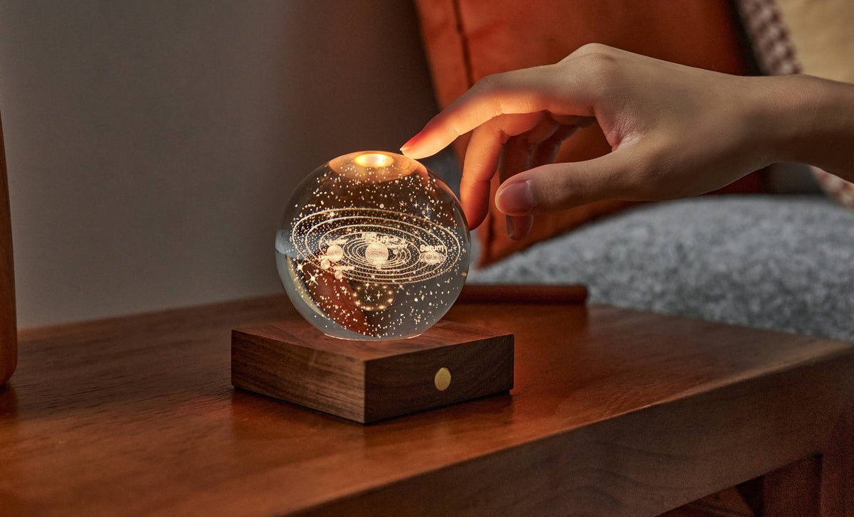 A hand reaching to touch the Gingko Amber Crystal Light, a glass sphere glowing warm on its walnut base