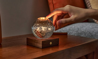 A hand reaching to touch the Gingko Amber Crystal Light, a glass sphere glowing warm on its walnut base