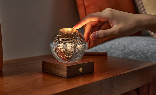 A hand reaching to touch the Gingko Amber Crystal Light, a glass sphere glowing warm on its walnut base