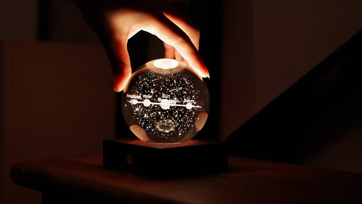 A hand resting on top of the lit crystal sphere on a dark desk beside a laptop