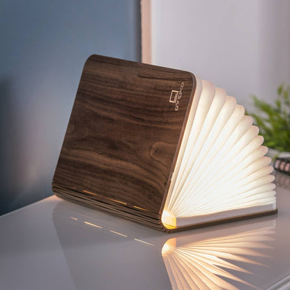 The walnut book light open on a table, its folded paper pages lit warm white
